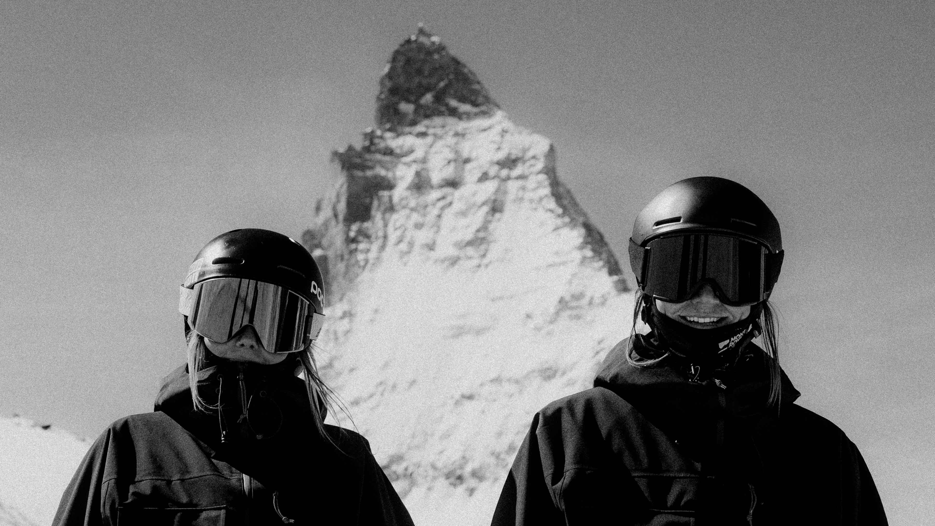 Two people in ski gear with goggles in front of a snow-covered mountain peak.