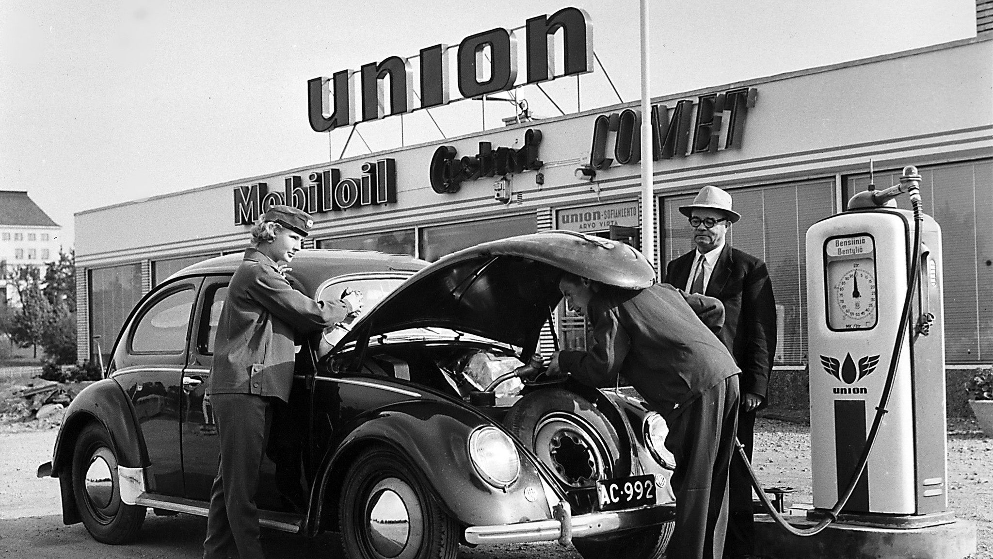 Vintage scene with a car at a gas station, featuring a Union Mobil Shell sign.
